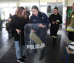 Policjantka pomaga dziewczynie chwycić tarczę, obok widoczny strażak.