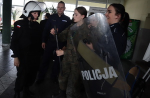 Dziewczyna ubrana w hełm obok inna trzyma policyjną pałkę, policjantka trzyma tarczę, za nimi stoi policjant.