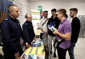 Dwóch umundurowanych policjantów stoi przy stoisku profilaktycznym i rozmawia z osobami zainteresowanymi służba w Policji.