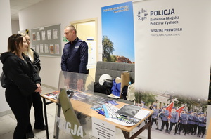 Policjant rozmawia z dwoma kobietami. Widoczny baner Komendy Miejskiej Policji w Tychach oraz stoisko profilaktyczne.