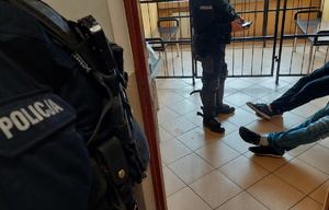 Policjant sprawdza na terminalu dane osoby, widoczne nogi dwóch zatrzymanych, na pierwszym planie kawałek munduru innego policjanta z napisem &quot;policja&quot;.