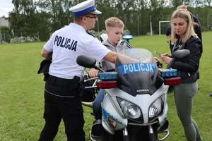 Umundurowany policjant Wydziału Ruchu Drogowego stoi przy motocyklu, na którym siedzi chłopak. Obok stoi kobieta.