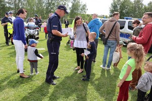 Umundurowany policjant podczas pikniku rodzinnego, trzyma kolorowanki, przed nim stoi chłopiec, obok niego kobieta. Widoczne inne osoby.