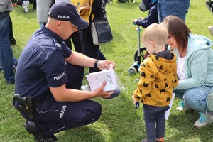 Umundurowany policjant kuca przy chłopcu, rozmawia z nim, trzyma kolorowanki. Obok chłopca kuca kobieta.