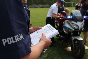 Napis &quot;Policja&quot; widoczny z koszuli munduru policjanta, który trzyma kolorowanki, w tle widoczny policjant i dziecko przy policyjnym motocyklu.