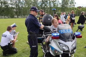 Jeden z policjantów stoi przy motocyklu, na którym siedzi dziewczynka, po lewej stronie policjant kuca przy chłopcu z założona policyjna czapką i daje mu opaskę odblaskową.