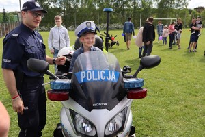 Umundurowany policjant stoi przy motocyklu, na którym siedzi dziewczynką z założoną policyjną czapką.