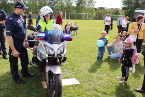 Policjant stoi przy motocyklu, na którym siedzi chłopiec w kasku, obok widoczni inni uczestnicy festynu.