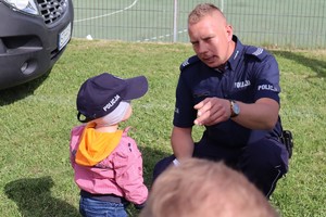 Umundurowany policjant kuca przy małym dziecku, które ma założona policyjną czapkę i rozmawia z nim pokazując coś palcem.