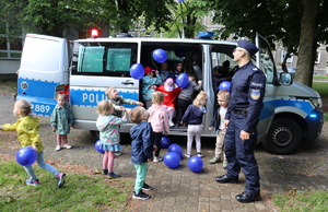 Umundurowany policjant stoi przy radiowozie w środku pojazdu oraz obok niego znajdują się dzieci, które odbijają balony.