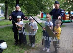 Policjanci stoją przy dzieciach, które trzymają pałki służbowe oraz tarcze.