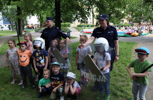 Policjant i policjantka stoją przy dzieciach, które ustawiły się do pamiątkowego zdjęcia trzymając pałki oraz policyjne tarcze.