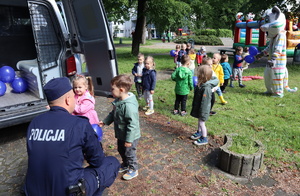 Policjant kuca przy dzieciach i rozmawia z nimi, obok policyjny radiowóz, dalej dzieci, pluszowa maskotka i dmuchany zamek.