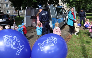 Na pierwszym planie balony z wizerunkiem policyjnej maskotki &quot;Sznupka&quot; dalej policjanci, dzieci i pani opiekunka przy policyjnym radiowozie.