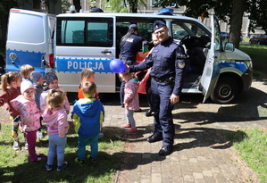 Umundurowany policjant trzyma balona, obok niego dzieci, dalej policjantka przy radiowozie.