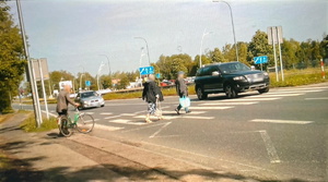 Na zdjęciu widoczne osoby przechodzące na przejściu dla pieszych oraz jeden samochód na przejściu i jeden samochód, który zatrzymał się przed przejściem.