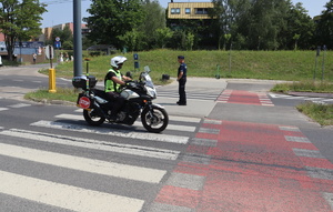 Na pasie rozdzielającym przejście dla pieszych stoi umundurowany policjant, po drodze jedzie motocyklista.