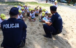 Policjanci kucają przodem do dzieci, które siedzą na piasku i rozmawiają z nimi o bezpieczeństwie nad wodą.