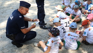 Policjant kuca przed dziećmi, które siedzą na piasku i rozmawia z chłopcem.