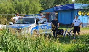 Policjanci stoją przy radiowozie, obok policjant oraz strażnik miejski z rowerami.