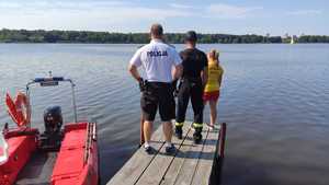 Zdjęcie przedstawiające policjanta, ratownika Wodnego Ochotniczego Pogotowia Ratunkowego oraz strażaka, którzy patrzą na jezioro stojąc na pomoście.