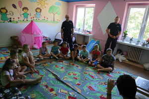 Dwóch umundurowanych policjantów stoi za siedząca na dywanie grupą dzieci.