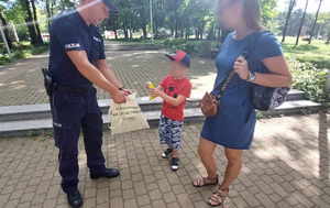 Umundurowany policjant wyciąga z torby piłkę chłopakowi, który stoi obok mamy.