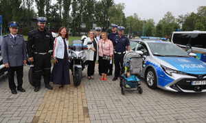 Zdjęcie grupowe policjantów z kobietami, widoczny policyjny motocykl oraz radiowóz.