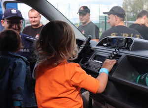 Dzieci siedzą w radiowozie obok stoi policjant, widoczni strażacy.