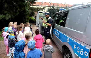 Umundurowany policjant z wydziału ruchu drogowego rozmawia z dziećmi, za nim policyjny radiowóz.