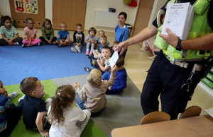 Dzieci siedzą na dywanie, umundurowany policjant rozdaje kolorowanki.
