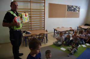 Umundurowany policjant trzyma w rekach podziękowania, widoczne dzieci na dywanie w przedszkolnej sali.
