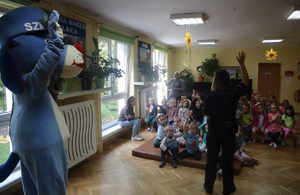Umundurowana policjanta stoi przed grupą dzieci z uniesioną ręką obok policyjna maskotka Sznupek z uniesioną łapą, przed nimi grupa dzieci z uniesionymi rękami, siedzących na ławkach oraz materacu.