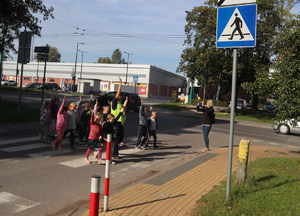 Policjantka w odblaskowej kamizelce unosi rękę do góry i przeprowadza przez przejście dla pieszych grupę przedszkolaków.