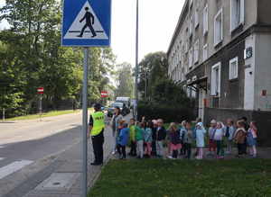 Umundurowana policjantka w odblaskowej kamizelce stoi z grupą dzieci przed przejściem dla pieszych.