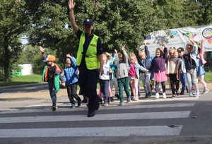 Policjantka w odblaskowej kamizelce idzie z uniesiona ręką przez przejście dla pieszych za nią idzie grupa dzieci.