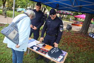 Stoisko promocyjne Komendy Miejskiej Policji w Tychach. Na zdjęciu dwóch umundurowanych policjantów oraz stolik z ulotkami, gadżetami i informatorami dla zainteresowanych. Przed stolikiem kobieta w starszym wieku, przyglądająca się rzeczom umieszczonym na stoliku.