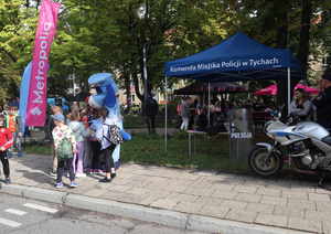 Stoisko promocyjne komendy Miejskiej Policji w Tychach. przed namiotem ustawiony motocykl policyjny. Po lewej stronie zdjęcia policyjna maskotka Sznupek i przytulające się do niej dzieci.