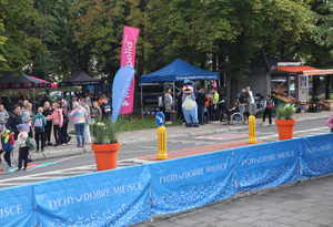 Fragment ulicy Czarnieckiego w Tychach. Pasy ruchu oddzielone plakatami z napisem &quot;Tychy dobre miejsce&quot;. W tle zdjęcia widoczne stanowisko promocyjne Komendy miejskiej Policji w Tychach, zgromadzeni uczestnicy imprezy oraz policyjna maskotka Sznupek.