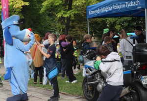 Stanowisko promocyjne Komendy Miejskiej Policji w Tychach. przed namiotem zaparkowany motocykl policyjny. Z boku namiotu stoi Sznupek, który przybija piątkę chłopcu.