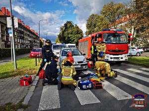 Zdjęcie przedstawiające strażaków, strażników miejskich oraz policjanta, którzy znajdują się na oznakowanym przejściu dla pieszych. Strażacy pochylają się nad noszami. W tle zdjęcia samochód osobowy, radiowóz Straży Miejskiej oraz wóz strażacki.