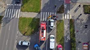 Zdjęcie wykonane przy użyciu drona. Widać na nim ulicę z oznakowanym przejściem dla pieszych. Na przejściu strażacy, strażnicy miejscy oraz policjant, stojacy wokół znajdujących się na przejściu noszy.