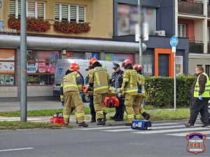 Zdjęcie przedstawiające strażaków, strażników miejskich oraz reportera. Strażacy oraz strażnicy przenoszą nosze z oznakowanego przejścia dla pieszych na chodnik.