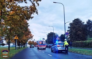 Policyjny radiowóz, policjant przy radiowozie, widoczna karetka pogotowia ratunkowego oraz inne pojazdy.