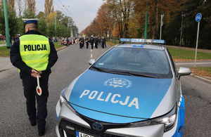 Policyjny radiowóz obok policjant z drogówki w kamizelce odblaskowej, dalej widoczni funkcjonariusze policyjnej orkiestry.