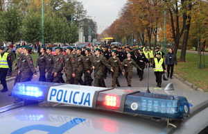 Napis &quot;Policja&quot; z sygnalizacji radiowozu, dalej maszerująca młodzież w mundurach po bokach osoby w odblaskowych kamizelkach.