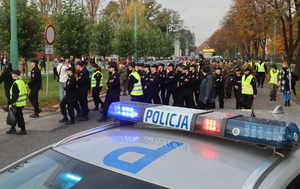 Na pierwszym planie widoczny napis &quot;Policja&quot; z radiowozu, dalej widoczni uczestnicy marszu, młodzież w mundurach, po bokach kolumny widoczne osoby w odblaskowych kamizelkach.