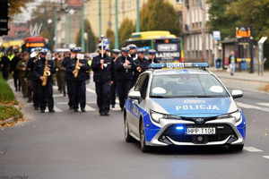 Policyjny radiowóz prowadzi przemarsz, na którym czele idą policjanci z orkiestry.