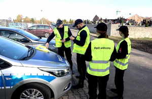 Policjanci oraz uczennice klasy mundurowej w żółtych kamizelkach, widoczny radiowóz.