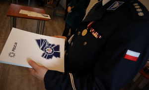 Policjant trzyma okładkę z logiem Policji widoczny jego mundur z odznaczeniami oraz pagony.